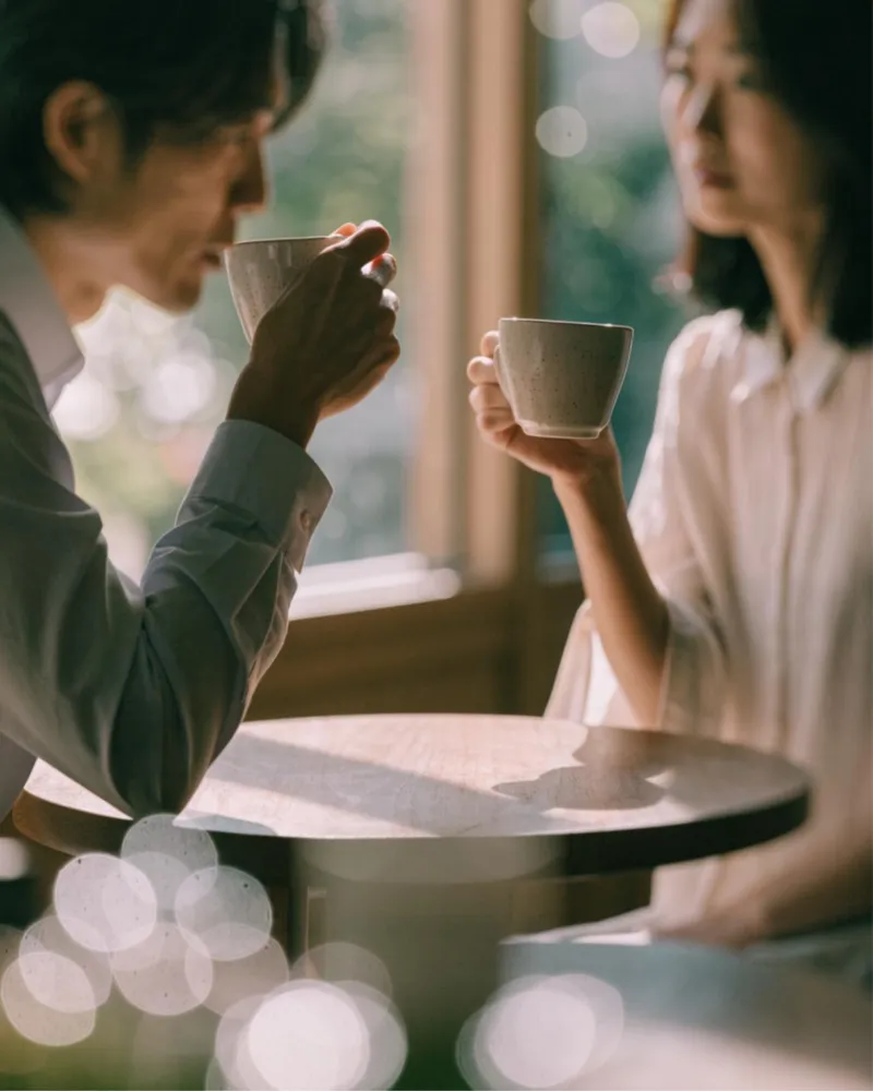 窓辺のカフェで向かい合い、コーヒーカップを手に話す二人の手元